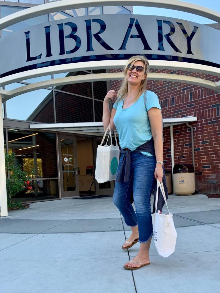 Writer as an adult, smiling with large book bags in front of the library signage.