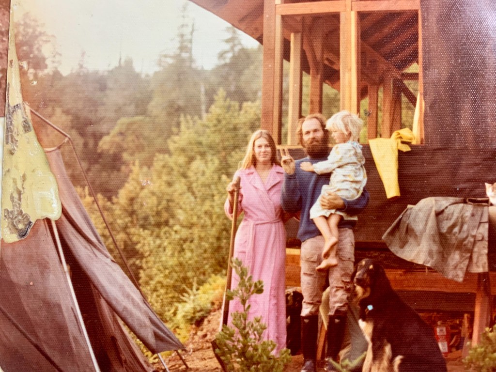 Writer as a chid (about age 4) poising with mom and dad in front of their in-progress a-frame cabin in  the classic style of Wood's famous painting: American Gothic. Her mom holds a shovel and dense trees are in the background.  