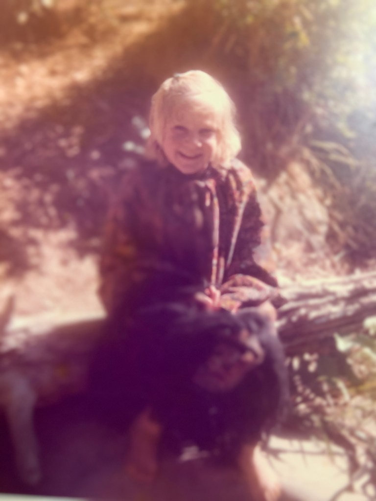 Writer at about age 5 sitting on a log smiling. She's wearing her mom's big poncho and the setting is in her family's outdoor "kitchen," which included only a camp stove and creek.