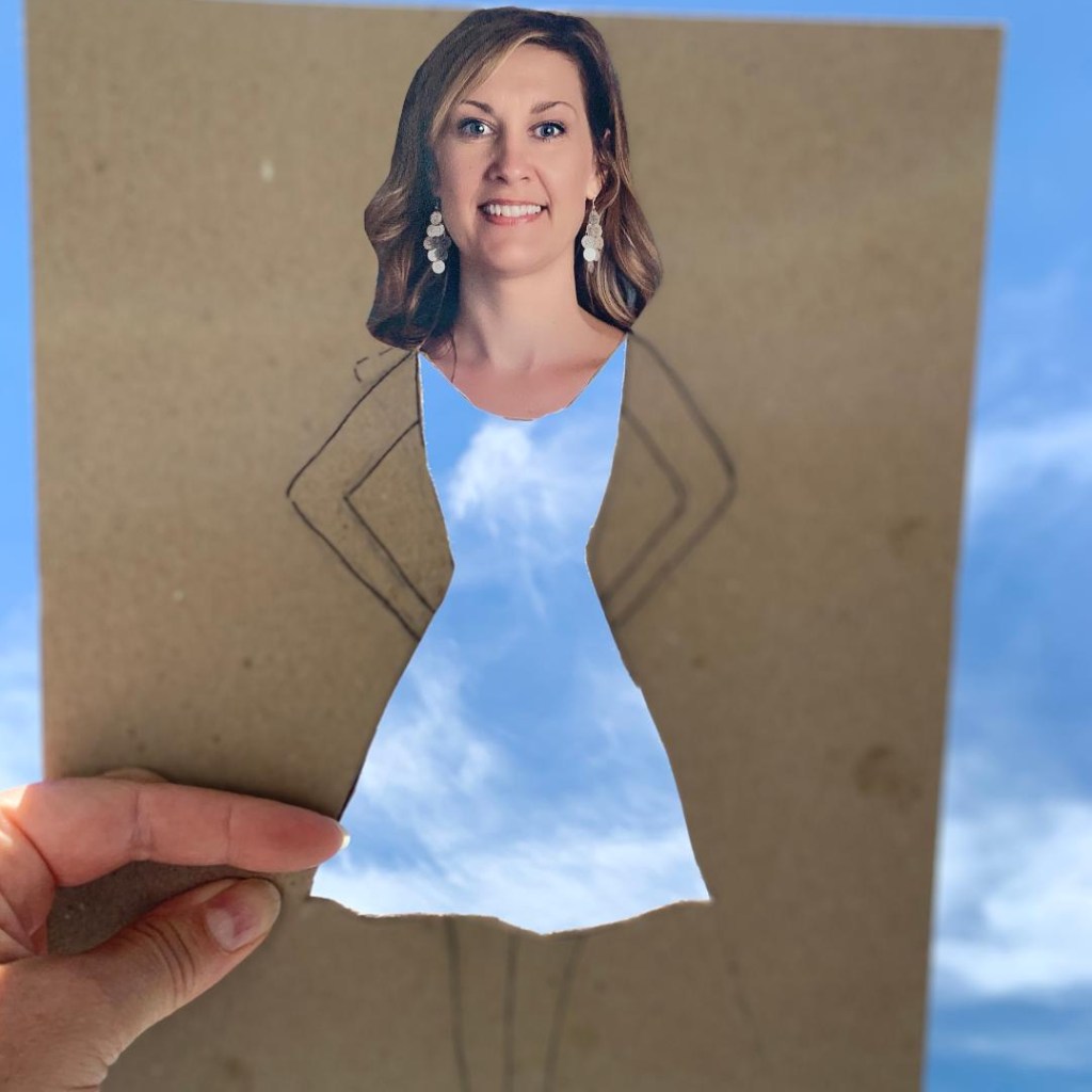 Writer holds a fun craft project up to let the sky show through the dress cut-out from a piece of cardboard that has writer's adult face cut and glued to top of dress with arms drawn in. 